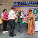 People on a school stage during a celebration at st Xaviers high school a man hands a bouquet to an elderly man while a student in a red uniform and a woman in a green sari stand nearby with a large banner in the background